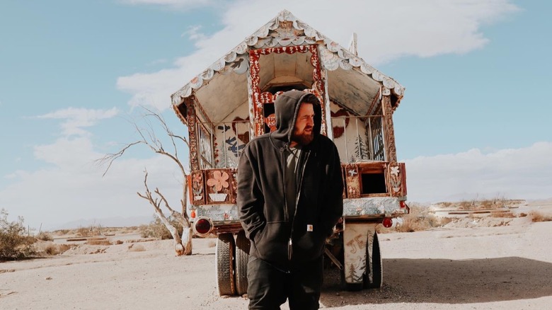 Lee DeWyze in a hoodie, standing in front of a house on wheels in the desert