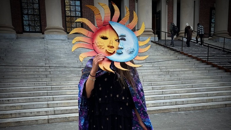 Jazz Jennings wears a cape and holds a sun and moon mask she made in front of her face.
