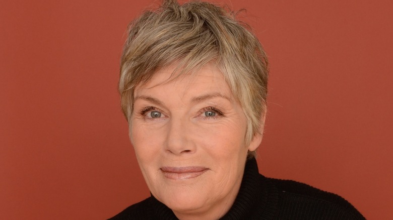 Kelly McGillis smiling wearing a black top in front of a brown background.