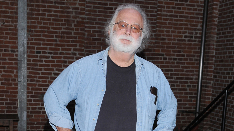 Brandon Maggart standing outside in a denim shirt and glasses with his hands on his hips.