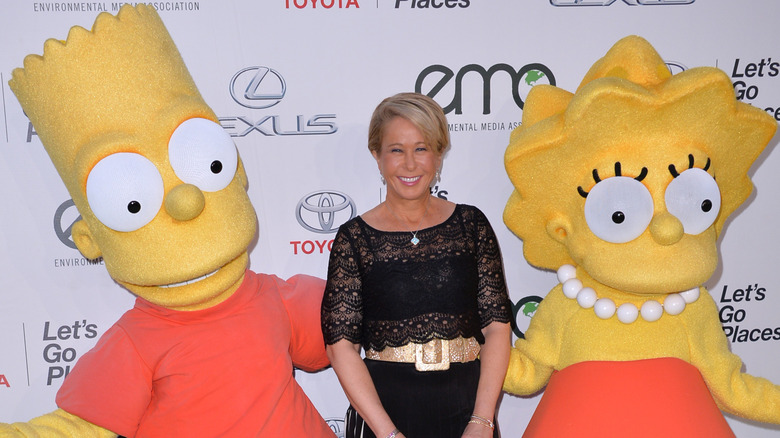 Yeardley Smith smiling and standing between two people wearing giant Bart and Lisa Simpson costumes.