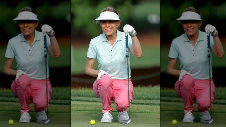 Vanna White kneels on a golf green in a visor holding a golf club smiling in a visor
