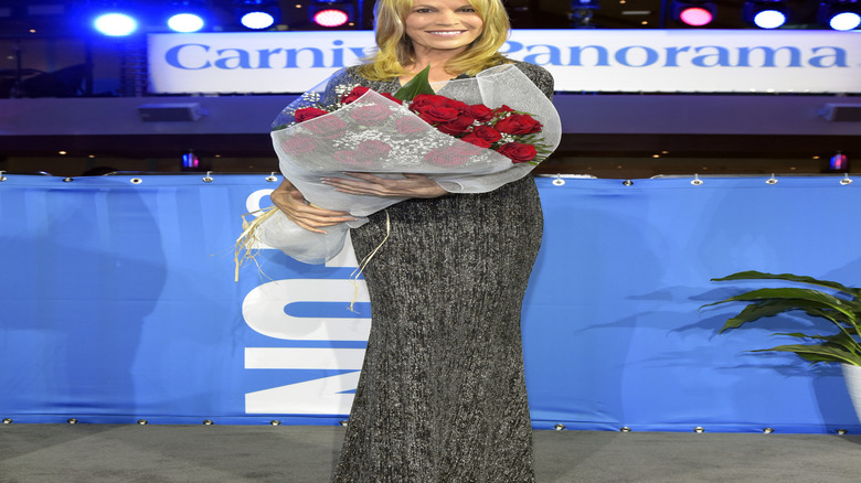Vanna White stands on a Carnival cruise ship smiling holding a bouquet of roses