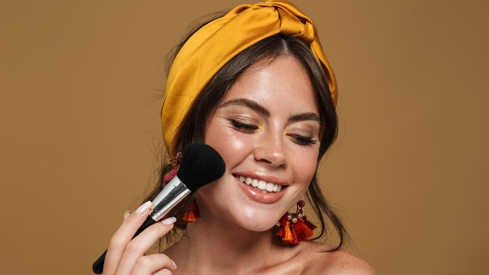 A woman is smiling while applying blush to her face.