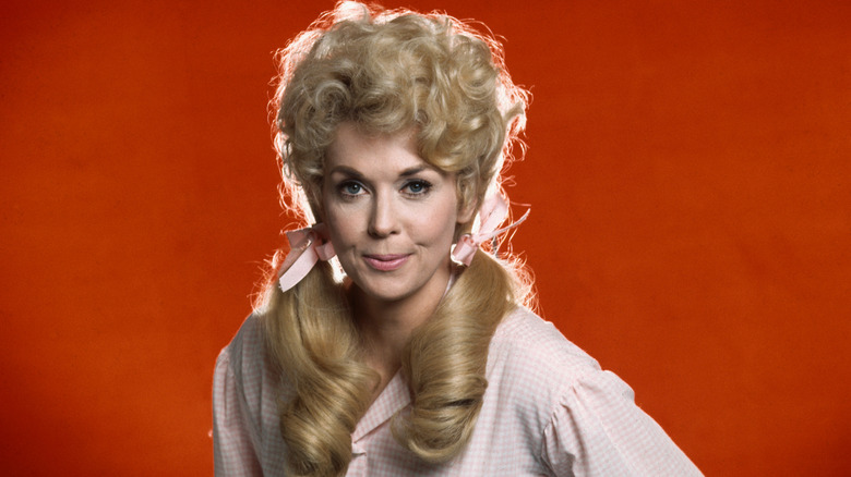 "The Beverly Hillbillies" actor Donna Douglas smiles in pigtails and a pink and white checkered shirt against a red background
