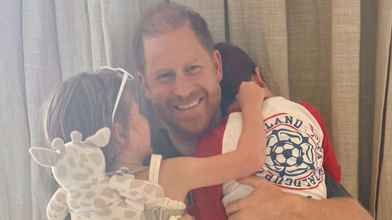A smiling Prince Harry hugging Princess Lilibet and Prince Archie