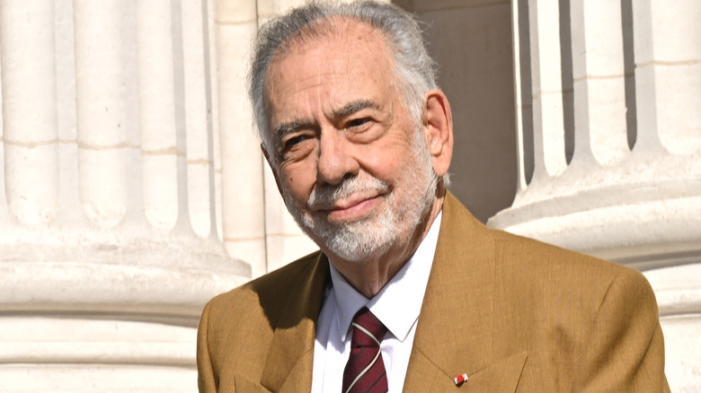 Francis Ford Coppola smiling in front of pillars in a brown suit