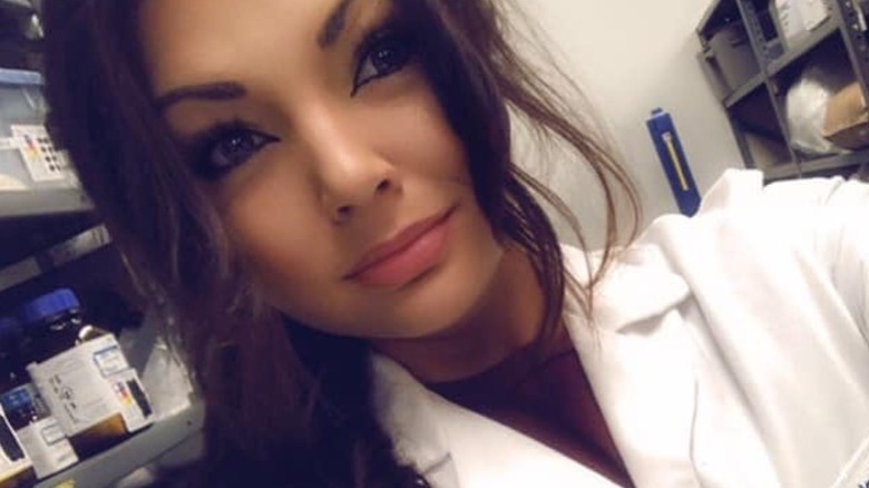 Closeup selfie of Stephanie Lebo in white lab coat in front of shelf with medications
