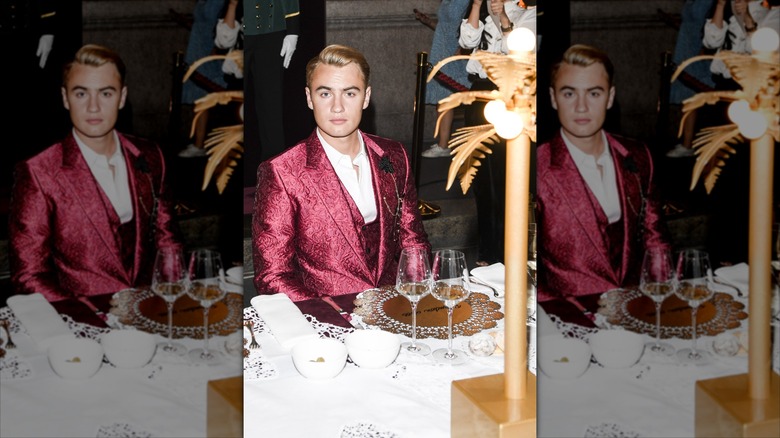 Brandon Thomas Lee in a red suit sitting in front of two empty wine glasses