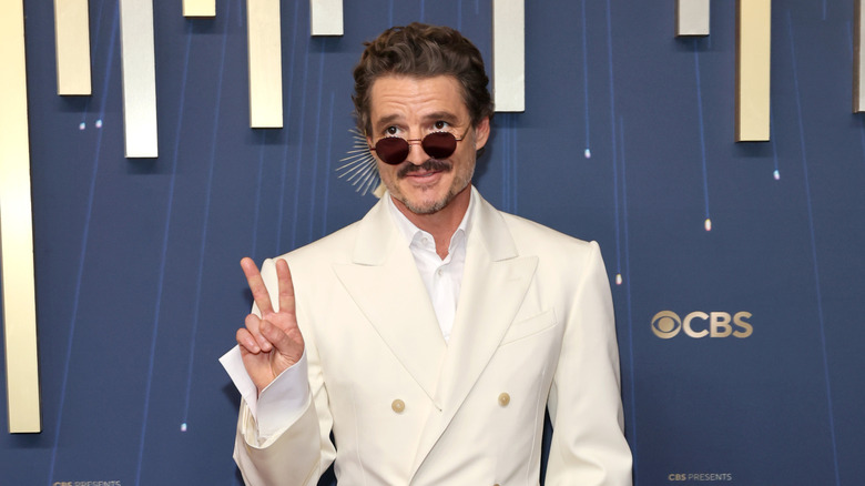 Pedro Pascal giving a peace sign while wearing sunglasses and a white suit