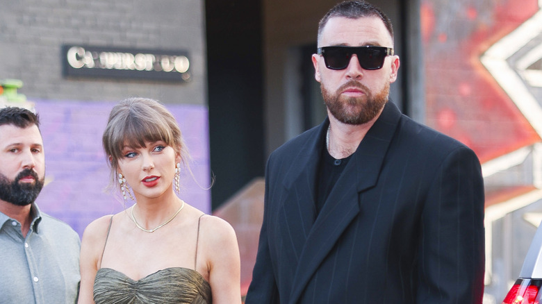 Taylor Swift wearing a chrome dress and Travis Kelce wearing a black suit and black sunglasses.