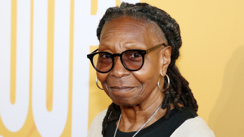 Whoopi Goldberg smiles at the camera in tinted glasses and gold hoops on the red carpet in 2026