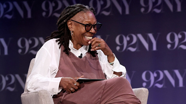 Whoopi Goldberg sitting while conducting an interview at a 92NY venue in a large white button up and mauve dress