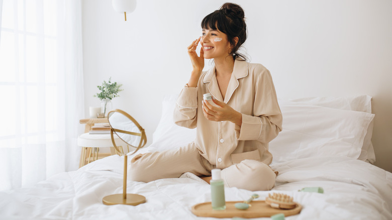 Woman applying skincare on bed smiling 