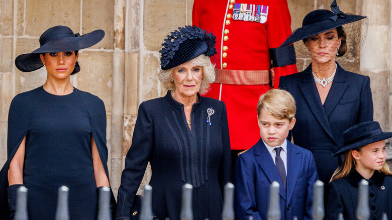 Meghan Markle, Queen Camilla, and Catherine, Princess of Wales with Prince George and Princess Charlotte all looking somber at Queen Elizabeth's funeral