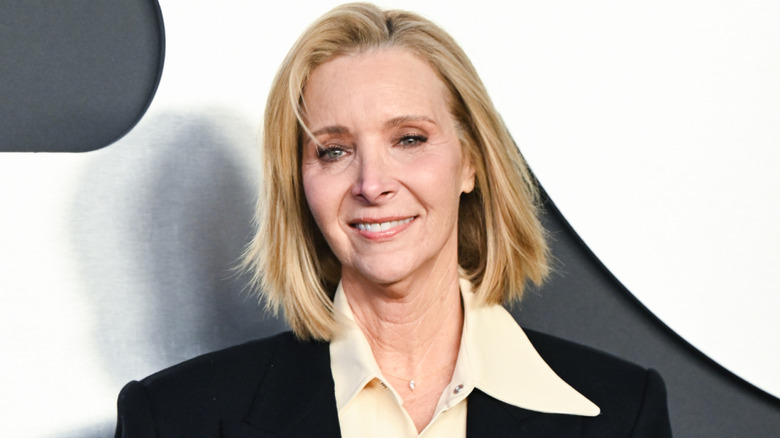 Lisa Kudrow smiling while posing at an event in a black blazer with a white blouse underneath