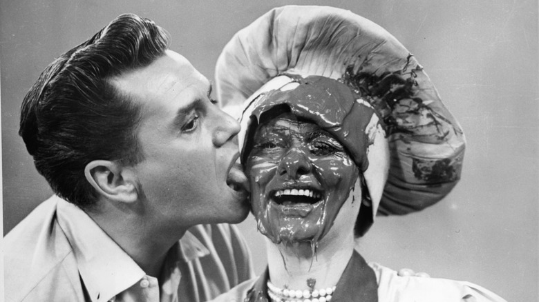 Black-and-white photo of Desi Arnaz licking Lucille Balls face while it's covered in chocolate and she laughs