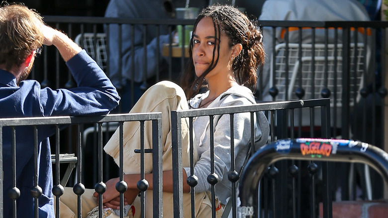 Malia Obama sitting in a chair, chewin on her lip, side-eying a camera.