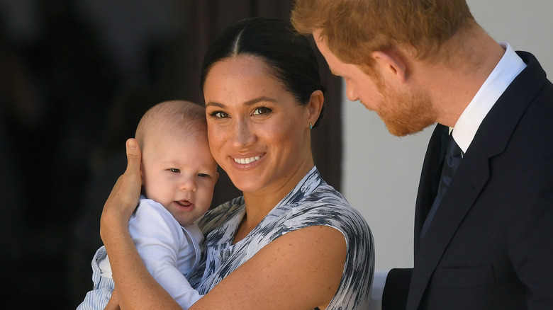 Meghan holding Archie, Harry hand on back