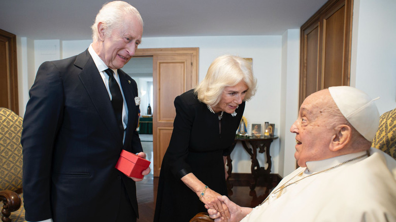 King Charles holding a box while Queen Camilla shakes hands with the Pope