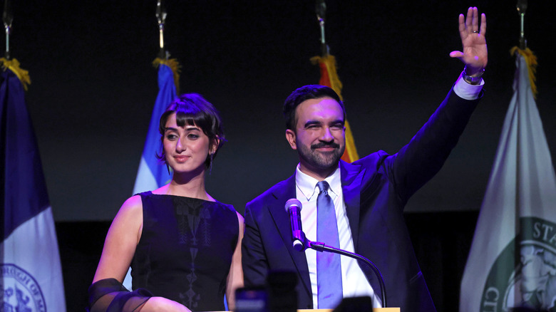 Zohran Mamdani waving while Rama Duwaji stands beside him onstage at an event