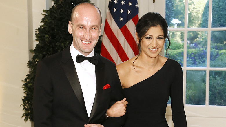 Stephen Miller in a tuxedo and Katie Miller in a black dress in front of a tree and the American flag