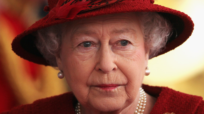 Queen Elizabeth II wearing a red hat, pearl earrings, and a pearl necklace looking off-camera