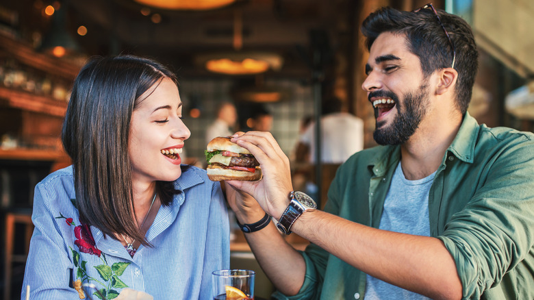 A couple on a date with a guy holding out his hamburger for a girl to take a bite