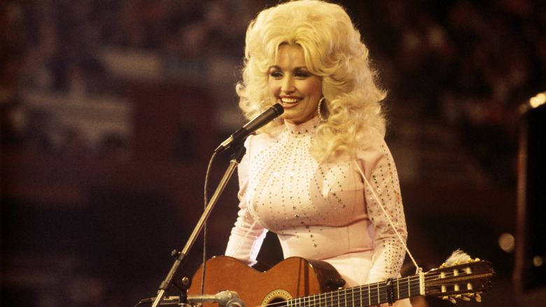 Dolly Parton performing in a baby pink embellished outfit and wavy blonde wig