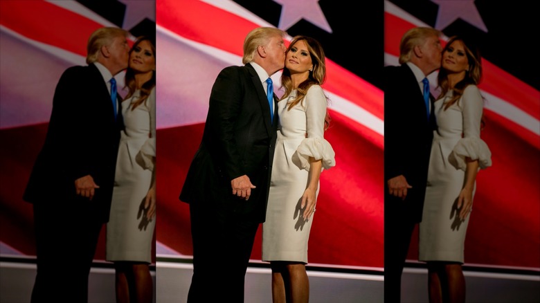 Donald Trump in a suit kissing Melania Trump on the cheek in front of a flag.