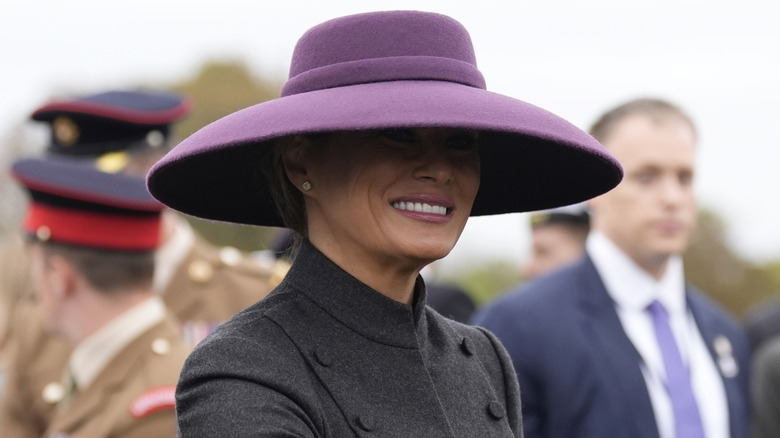Melania Trump smiling under the shadow of the wide brim of her purple hat.