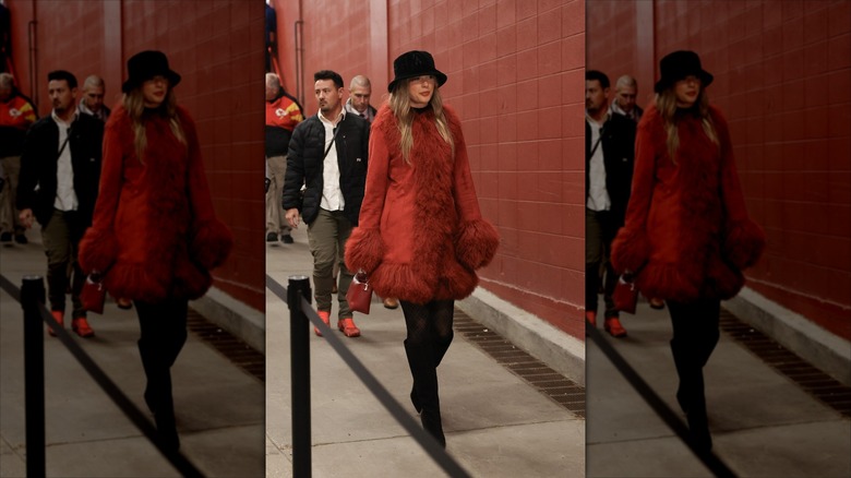 Taylor Swift in a furry red jacket and bucket hat, walking through a red tunnel.