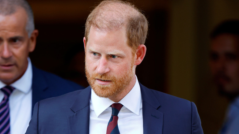 Prince Harry looking serious in a navy suit with a red and navy tie and white shirt