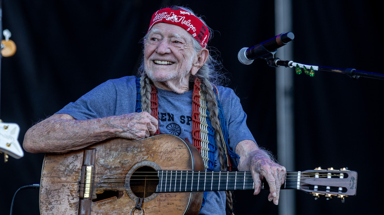 Willie Nelson in blue shirt and red bandana sitting in front of mic holding guitar