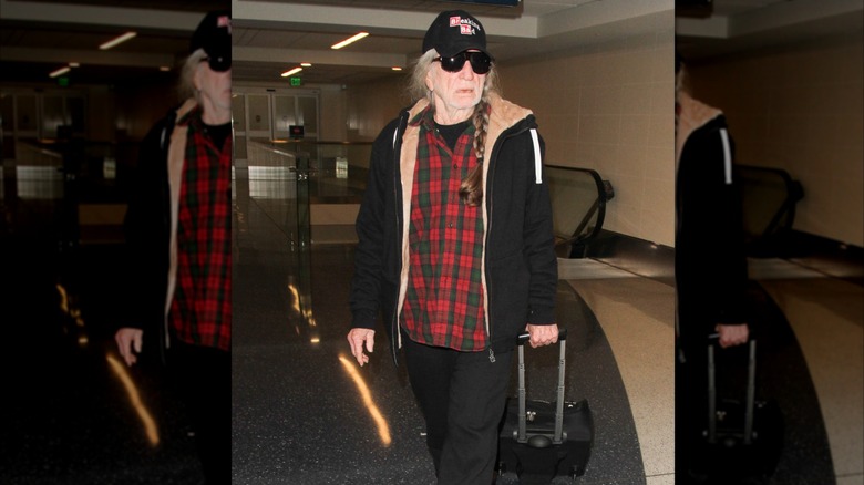 Willie Nelson wearing plaid shirt, ball cap, and sunglasses while wheeling luggage