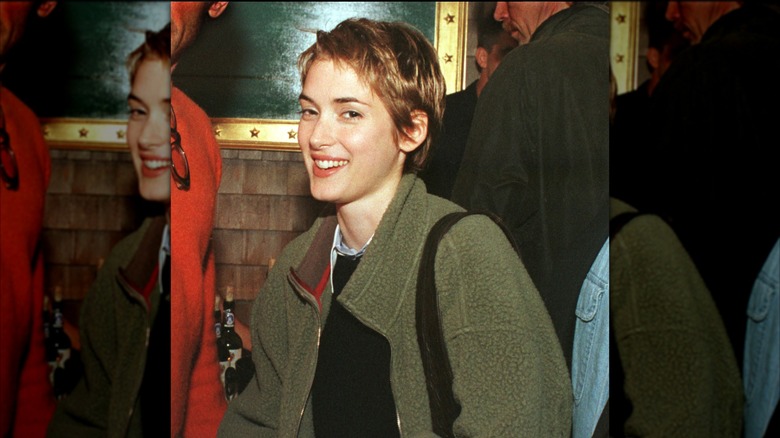 Winona Ryder smiles in a black shirt and olvie green jacket