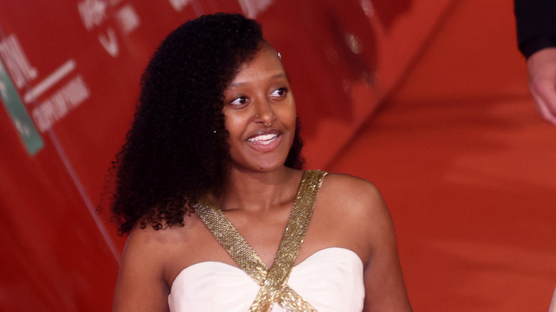 Zahara Jolie-Pitt grins while on the red carpet in a white dress with medium curly hair
