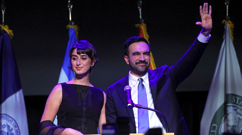 Zohran Mamdani and Rama Mamdani lit in a blue light, standing behind a podium.