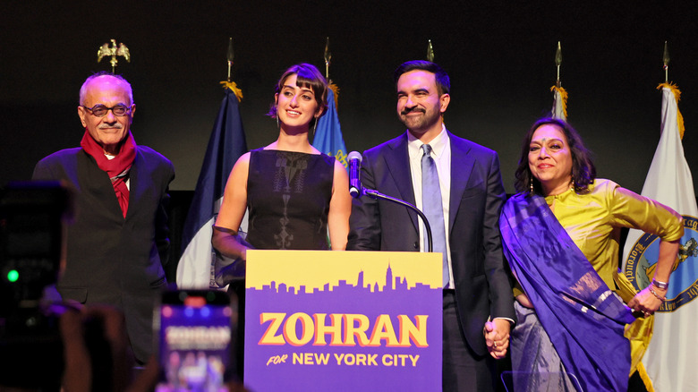 Mahmood Mamdani, Rama Duwaji, Zohran Mamdani, and Mira Nair standing at podium in front of flags