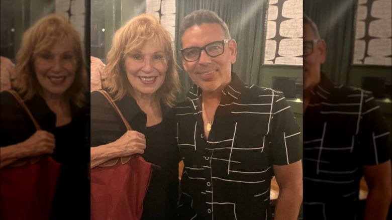 Joy Behar holding a large red purse on her shoulder and posing next to a man