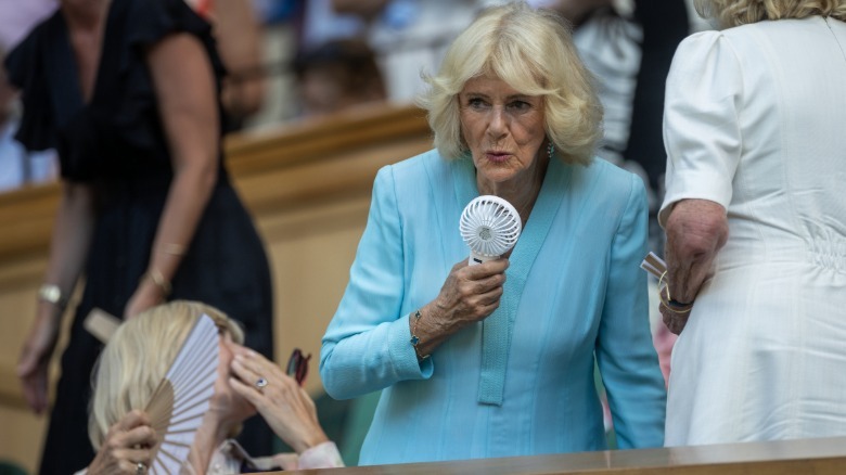 Queen Camilla using a white handheld fan to cool herself at Wimbledon